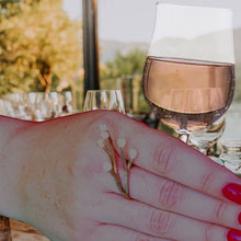 Load image into Gallery viewer, White woman wearing vermeil yellow gold ring with a twig design, handling a glass with a pink orange drink, table set for meal with guests in the background